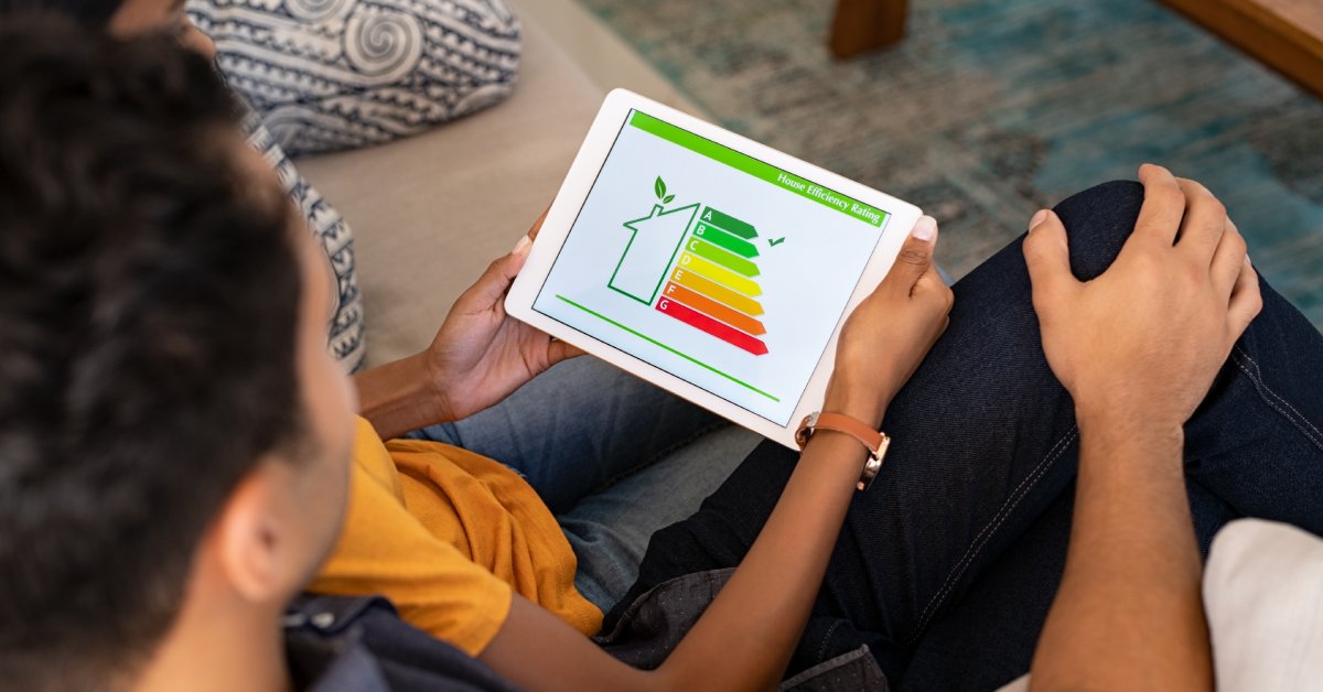 Two people sitting on a couch look at a tablet with a graphic of a home, a leaf, and different bars of green and red.