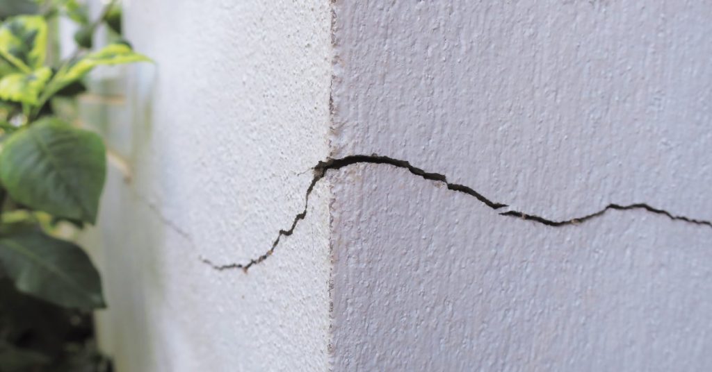 A horizontal, curved crack on an exterior corner of a building's white wall. Green plants are in the background.