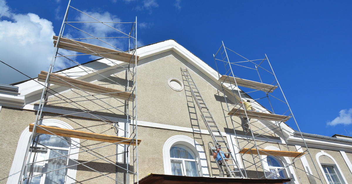 A building with scaffolding in front of it. There is a blue sky and a man on the scaffolding holding a ladder.