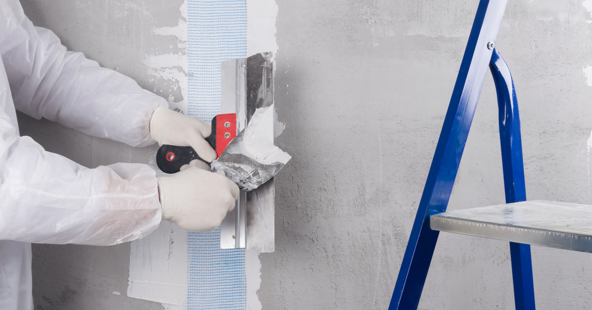 A person wearing white gloves applying plaster to a gray wall. There is a ladder to the right of the person.