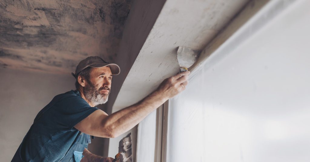 A worker in a ball cap uses a trowel to level out plaster on the underside of an eave, next to a large window.