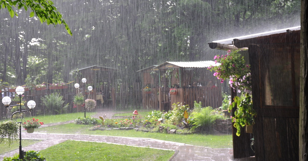 A small courtyard or garden area with wooden structures, greenery, and lampposts. It is pouring rain.