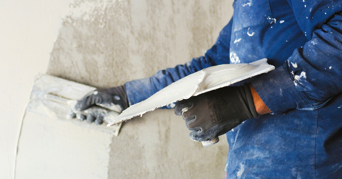 A worker wearing gloves and a work shirt places plaster on a wall and smooths it out with two different trowels.