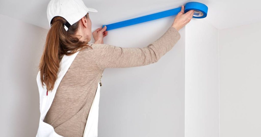 A person wearing overalls and a white hat uses blue painter's tape to block off areas of a large white wall.