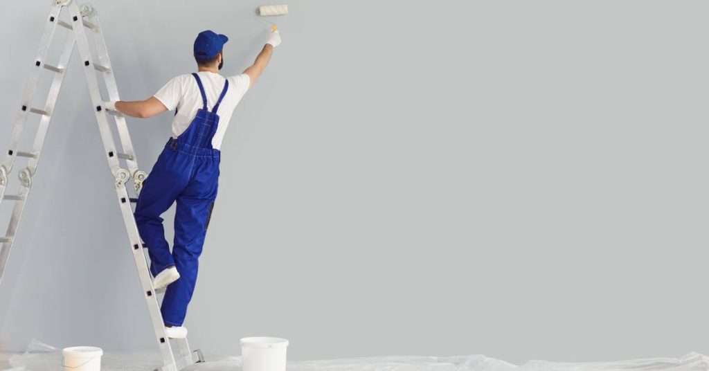 A worker in blue overalls holds onto a ladder with one hand and uses their other hand to paint a large wall.