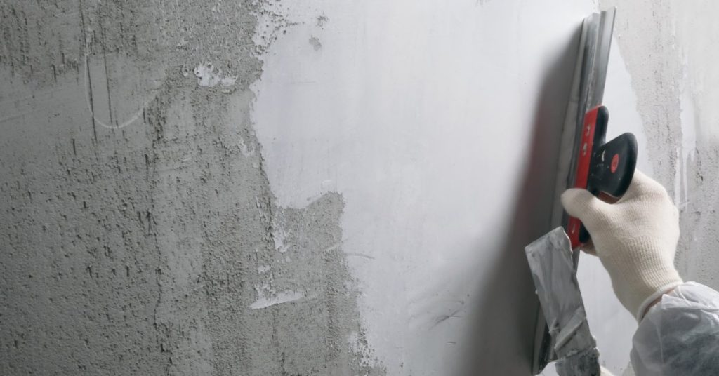 A worker wearing gloves and a protective jacket uses tools to work on the plaster of a home, flattening it out.