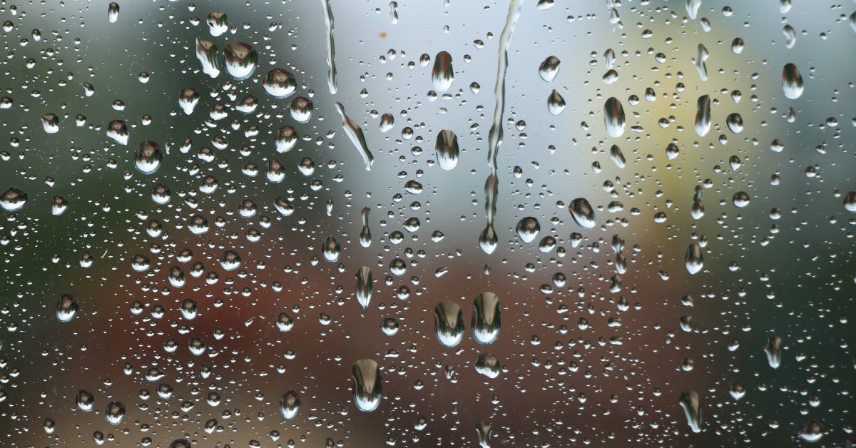 A close-up of different-sized rain drops on a window with blurry colors behind it, like blue, yellow, and red.