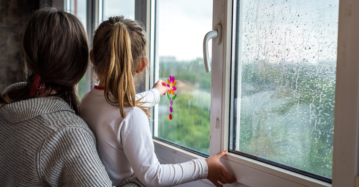 A woman holding a young girl stands by a window with raindrops on it, looking at the rain and greenery outside.